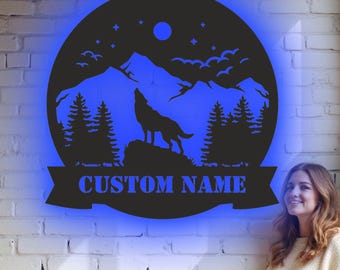 Op de afbeelding: Zwarte metalen wanddecoratie met een silhouet van een huilende wolf tegen een berg- en boslandschap, verlicht door blauwe LED-verlichting. De banner hieronder luidt "CUSTOM NAME".