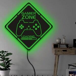 Może przedstawiać: Czarno-zielony znak strefy gier z podświetleniem zielonym. Znak zawiera kontroler do gier wideo i tekst "GAMER ZONE" oraz "LOADING..."