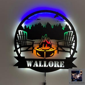 Pode incluir: Uma decoração de parede de metal preto com uma silhueta de uma fogueira, duas cadeiras e uma floresta. A decoração é iluminada com luzes LED multicoloridas e tem o nome "WALLORE" em uma faixa abaixo da imagem.