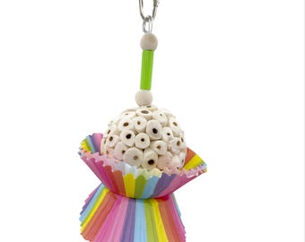 May include: A bird toy with a white, spherical body made of small, hollow cylinders, set in a colorful, rainbow-striped paper cup. The toy hangs from a metal clip and features a green and wooden bead accent, and a silver bell at the bottom.