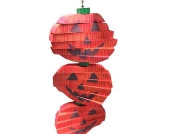 May include: A hanging Halloween decoration featuring three stacked, orange wooden jack-o'-lanterns with carved faces. The pumpkins are connected by green wooden stems and a silver chain. A small silver bell hangs at the bottom.