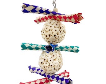 May include: A colorful bird toy with a metal clip for hanging. The toy features two white, textured spheres, woven blue, red, and teal accents, and a small green and blue ring. The toy is designed for birds to play with.