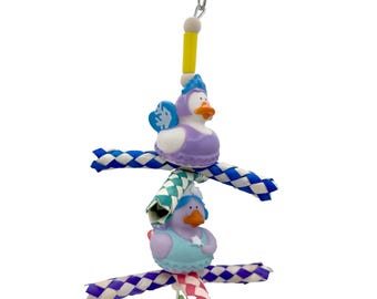 May include: A bird toy featuring two duck figurines in purple and blue outfits, connected by braided rope. The toy includes a metal clip and colorful accents, designed for avian enrichment and play.