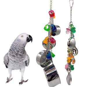 Puede incluir: Un loro gris está de pie cerca de dos juguetes colgantes para pájaros. Los juguetes están hechos de tazas y cucharas medidoras de acero inoxidable, con cuentas y abalorios de plástico de colores. Los juguetes están suspendidos de cadenas de metal.