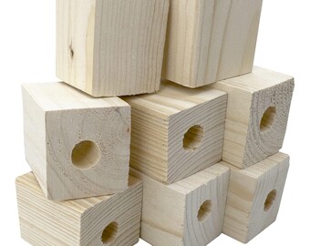 May include: A stack of unfinished, light-colored wooden blocks. Each block is a cube shape, with a hole drilled through one side. The wood grain is visible on the surface of the blocks. The blocks are stacked in a haphazard arrangement.