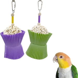 Puede incluir: Dos juguetes para pájaros con forma de magdalenas, uno morado y otro verde, colgando de clips plateados. Las coberturas de las magdalenas están hechas de pequeños trozos cilíndricos blancos. Estos juguetes están diseñados para aves.