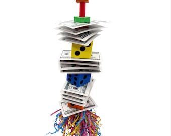 May include: A colorful bird toy featuring wooden blocks, dice, and stacks of paper currency. The toy is topped with a metal clip and has a fringe of colorful paper strips at the bottom. The dice are yellow and blue with black dots.