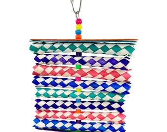 May include: A colorful bird toy with woven, layered sections in various colors, including teal, purple, pink, and blue. The toy is suspended by a metal clip and features small, colorful beads. A small purple ring hangs from the bottom.