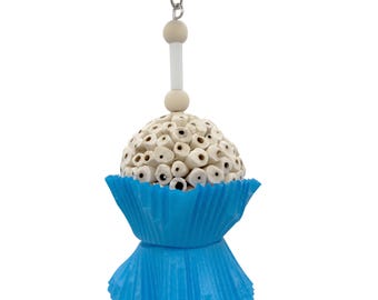 May include: A bird toy with a white, textured sphere made of small, cylindrical pieces, resembling a cupcake. It sits atop a bright blue, pleated paper cup. The toy hangs from a metal chain and has wooden beads.