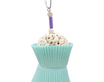 May include: A bird toy shaped like a cupcake, featuring a light blue paper cup base and a white, textured top. A purple tube and wooden beads hang from a metal clip at the top. The toy is designed for birds to play with.