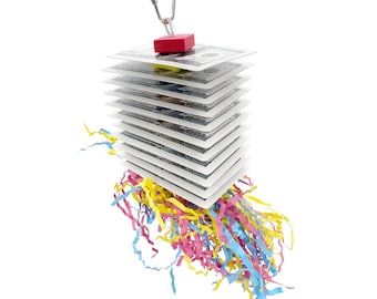 May include: A bird toy featuring a stack of square cards with a money design, topped with a red wooden cube and suspended by a metal clip. Colorful shredded paper in yellow, pink, and blue hangs from the bottom.
