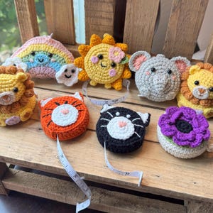 May include: A selection of colourful, crocheted tape measures in the shapes of a rainbow, sun, lion, elephant, tiger, cat, and flower. Each tape measure has a unique design and is made of yarn. The tape measures are displayed on a wooden surface.