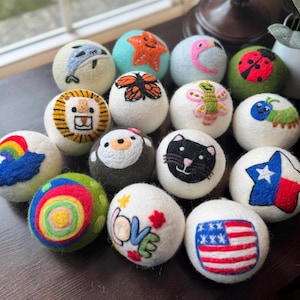 May include: A collection of white felt balls, each adorned with colorful embroidered designs. The designs include a dolphin, starfish, butterfly, ladybug, lion, cat, rainbow, and the word "love". The balls are arranged on a dark brown surface.