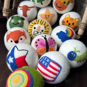 May include: A collection of white felted wool balls featuring various needle-felted designs including a green tree, a giraffe, a longhorn, a starfish, a cat, a frog, a lion, a fox, a butterfly, a rainbow, a Texas flag, a caterpillar, and a US flag.