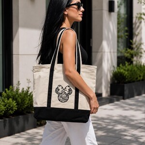 May include: A beige tote bag with black accents and handles. The bag features a black decorative design on the front. The bottom of the bag is black. The bag is carried by a person.