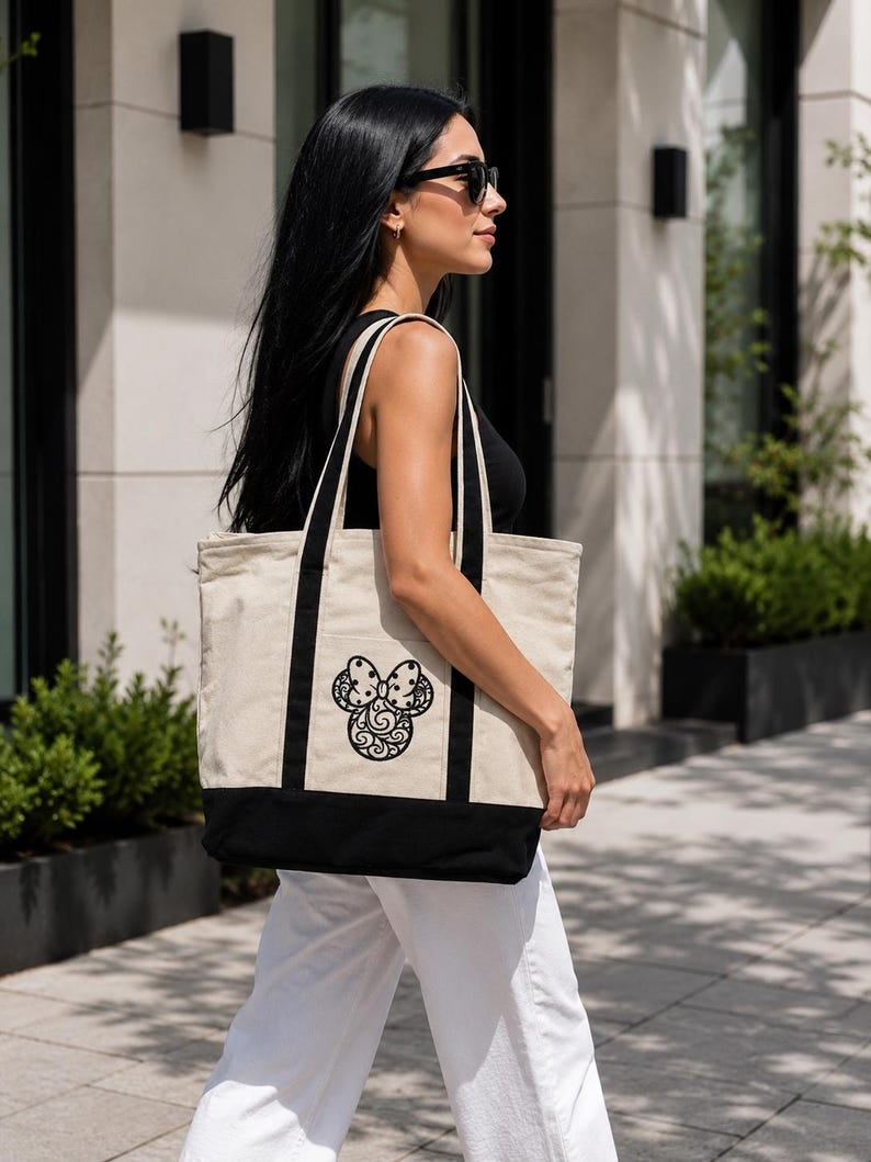 May include: A beige tote bag with black accents and handles. The bag features a black decorative design on the front. The bottom of the bag is black. The bag is carried by a person.