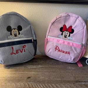 Puede incluir: Dos mochilas de seersucker con personajes de Disney bordados. La bolsa de rayas azules y blancas muestra a Mickey Mouse y el nombre Levi en rojo. La bolsa de rayas rosas y blancas presenta a Minnie Mouse y el nombre Parker en rojo.