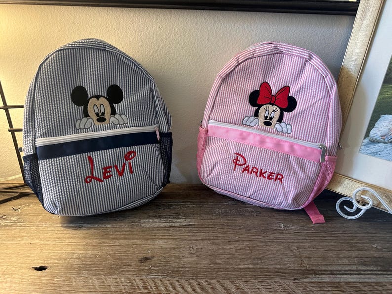Puede incluir: Dos mochilas de seersucker con personajes de Disney bordados. La bolsa de rayas azules y blancas muestra a Mickey Mouse y el nombre Levi en rojo. La bolsa de rayas rosas y blancas presenta a Minnie Mouse y el nombre Parker en rojo.