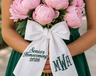 May include: A bouquet of light pink peony flowers with green leaves, tied with a white fabric ribbon. The ribbon features dark green embroidered text that reads Senior Homecoming 2026 on one side and the monogram MWA on the other side.