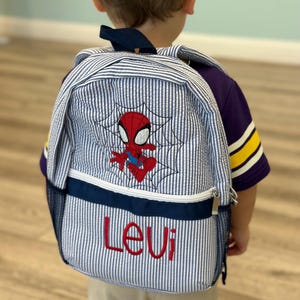May include: A blue and white striped backpack featuring an embroidered Spider-Man design and the name "Levi" in red. The backpack has a navy blue trim and a mesh side pocket. The straps are navy blue.