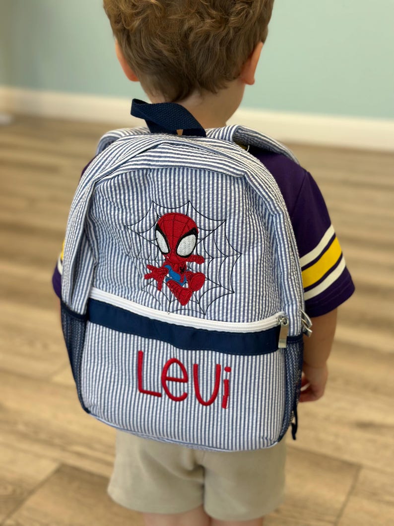 Op de afbeelding: Een blauw-wit gestreepte rugzak met een geborduurd Spider-Man-ontwerp en de naam "Levi" in het rood. De rugzak heeft een marineblauwe rand en een zijvak van gaas. De bandjes zijn marineblauw.