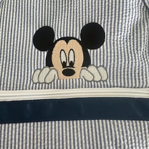 Puede incluir: Una mochila de tela seersucker azul y blanca con un bordado de Mickey Mouse asom&aacute;ndose sobre una cremallera blanca horizontal y un borde de cinta azul marino. El dise&ntilde;o incluye las ic&oacute;nicas orejas negras y el rostro del personaje.