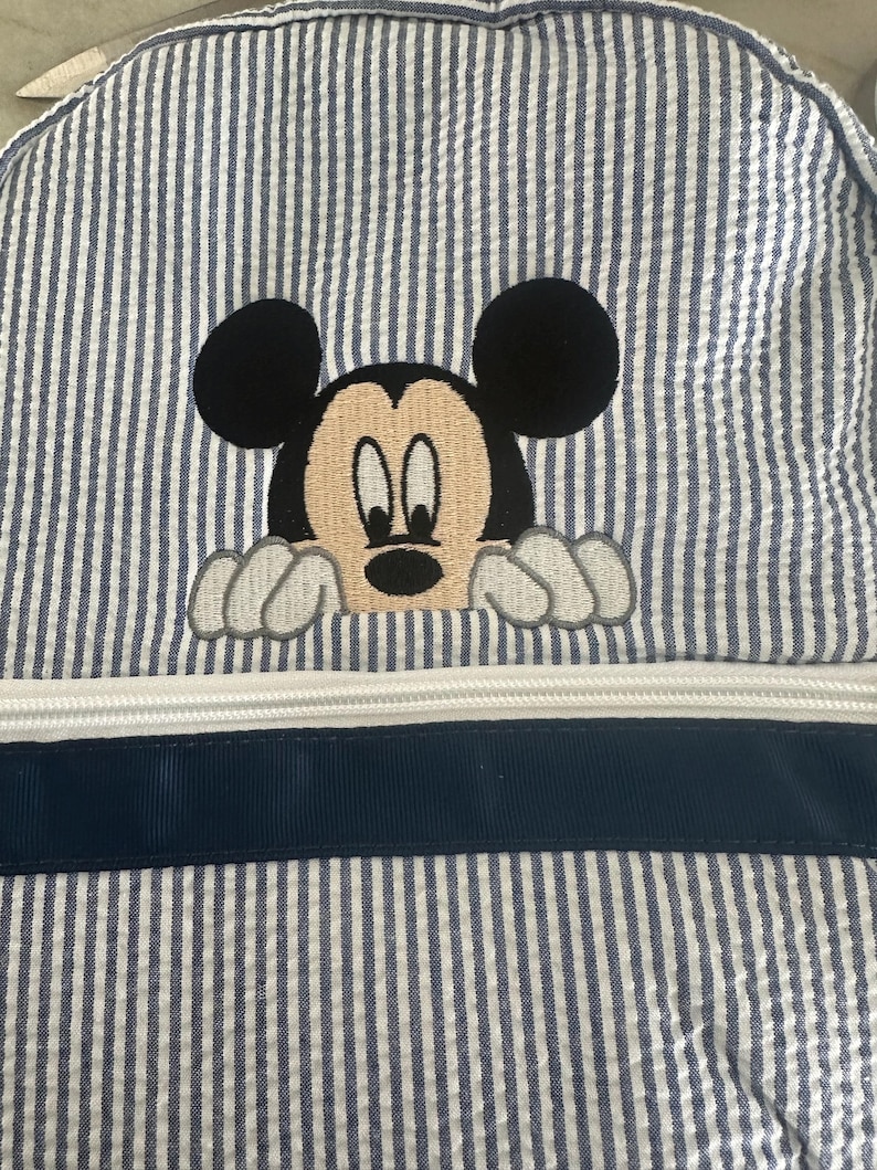 Puede incluir: Una mochila de tela seersucker azul y blanca con un bordado de Mickey Mouse asom&aacute;ndose sobre una cremallera blanca horizontal y un borde de cinta azul marino. El dise&ntilde;o incluye las ic&oacute;nicas orejas negras y el rostro del personaje.
