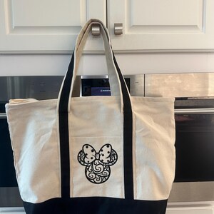 May include: A beige canvas tote bag with black accents and a black decorative bow design. The bag has a black bottom, black handles, and a front pocket. The bag is hanging in front of a white cabinet.