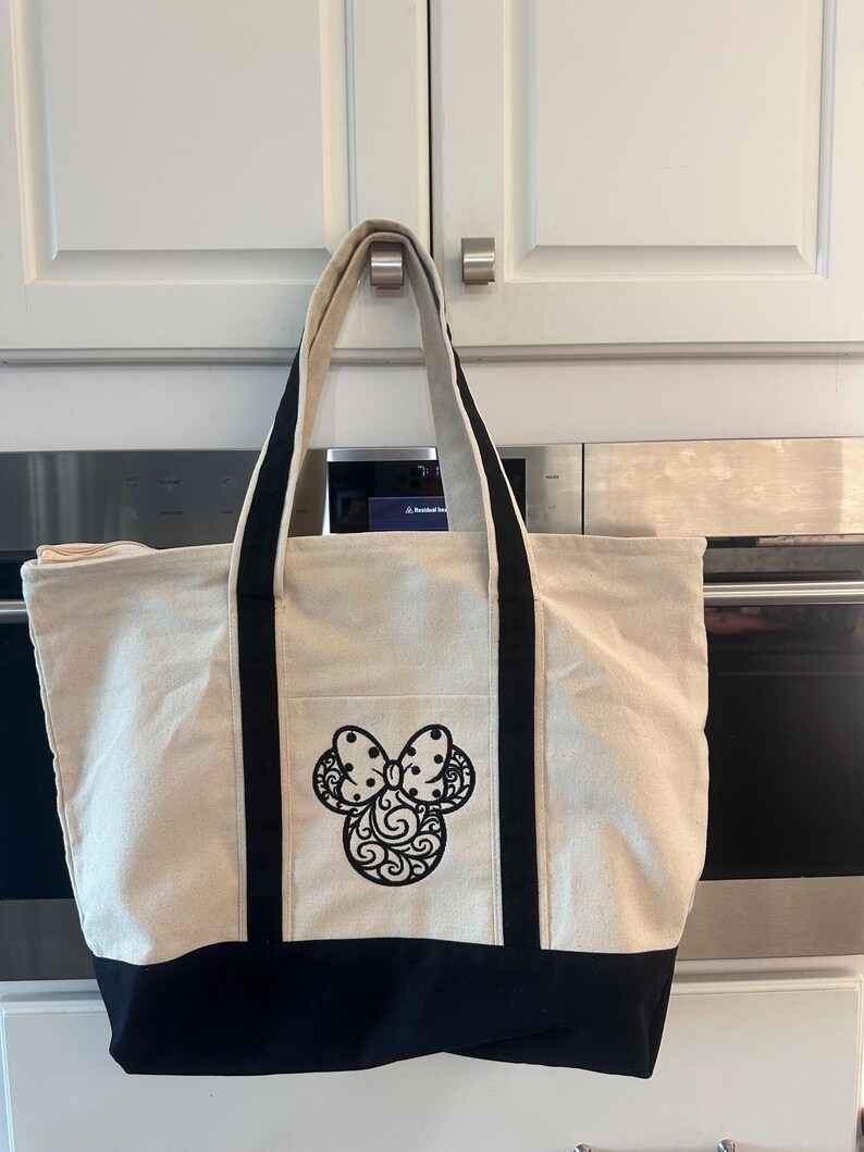 May include: A beige canvas tote bag with black accents and a black decorative bow design. The bag has a black bottom, black handles, and a front pocket. The bag is hanging in front of a white cabinet.