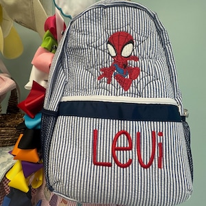 Op de afbeelding: Een blauw-wit gestreepte rugzak met een geborduurd Spider-Man-ontwerp en de naam "Levi" in het rood. De rugzak heeft een marineblauwe rand en een zijvak van gaas. Het ontwerp bevat een spinnenweb.