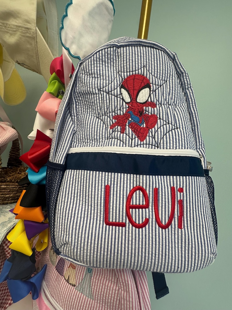 Op de afbeelding: Een blauw-wit gestreepte rugzak met een geborduurd Spider-Man-ontwerp en de naam "Levi" in het rood. De rugzak heeft een marineblauwe rand en een zijvak van gaas. Het ontwerp bevat een spinnenweb.