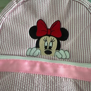 Puede incluir: Una mochila de seersucker rosa y blanca con un bordado de Minnie Mouse con un lazo rojo. La bolsa incluye un adorno de cinta rosa claro horizontal en la parte delantera, situado sobre el cierre de cremallera blanco del compartimento.