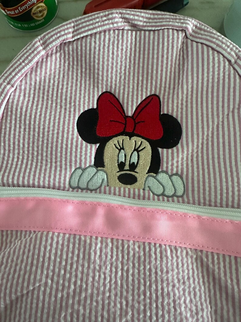 Puede incluir: Una mochila de seersucker rosa y blanca con un bordado de Minnie Mouse con un lazo rojo. La bolsa incluye un adorno de cinta rosa claro horizontal en la parte delantera, situado sobre el cierre de cremallera blanco del compartimento.