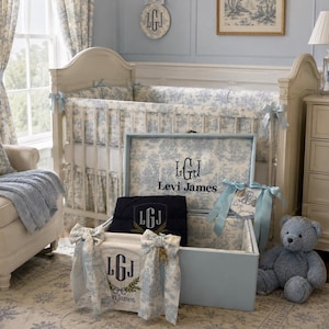 May include: A light blue storage trunk featuring a toile pattern interior, personalized with the monogram LGJ and the name Levi James. Inside, a dark navy blanket with an embroidered LGJ crest sits alongside a white fabric accessory with bows.