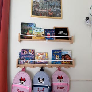 Puede incluir: Dos estantes de pared de madera clara con libros infantiles y un tablero de letras negro con el texto 3 DAYS UNTIL DISNEY WORLD. Debajo, tres mochilas peque&ntilde;as cuelgan de ganchos: dos bolsos de Minnie Mouse a rayas rosas y uno de Mickey Mouse a rayas azules.