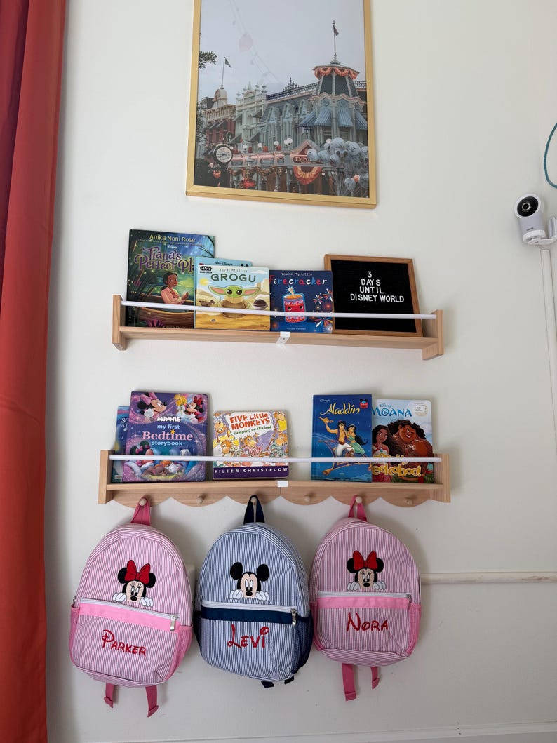Puede incluir: Dos estantes de pared de madera clara con libros infantiles y un tablero de letras negro con el texto 3 DAYS UNTIL DISNEY WORLD. Debajo, tres mochilas peque&ntilde;as cuelgan de ganchos: dos bolsos de Minnie Mouse a rayas rosas y uno de Mickey Mouse a rayas azules.