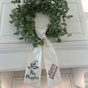 May include: A white fabric wreath with the text "No Muggles" and "Wizards Welcome" embroidered on it. The wreath is decorated with a green leafy garland.