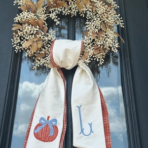May include: A fall-themed wreath with a cream-colored ribbon and red plaid trim. The ribbon is embroidered with a red pumpkin and blue bow, and the letter "J". The wreath is made of brown leaves and white berries.