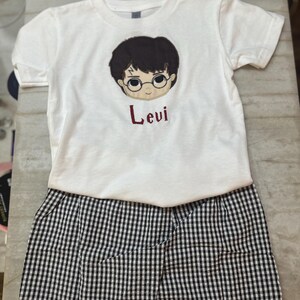 May include: A white t-shirt and black and white checkered shorts set. The t-shirt features a cartoon illustration of a boy with glasses and the name "Levi" in red. The shorts are a classic gingham pattern.