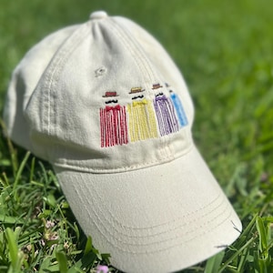 May include: A beige baseball cap with an embroidered design of four figures in hats and bow ties, each with a different colored vertical stripe pattern: red, yellow, purple, and blue. The cap is sitting on green grass.
