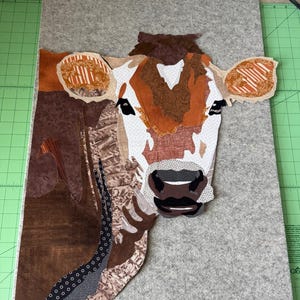 May include: A fabric art piece depicting a cow's head. The cow's face is white with brown and orange patches. The ears have orange and white striped patterns. The background is a gray felt-like material.