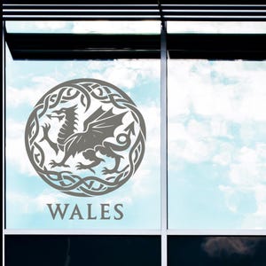May include: A grey circular decal featuring a dragon within a Celtic knot design. The word "WALES" is printed below the emblem. The decal is affixed to a window with a blue sky visible in the background.