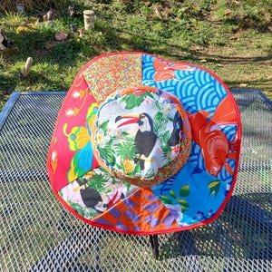 May include: A large, red, white and blue sun hat with a toucan print. The hat is made of patchwork fabric and has a wide brim.