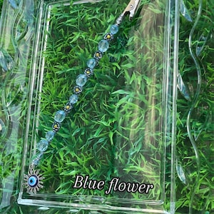 May include: A blue and gold beaded clip with an evil eye charm. The clip is on a green background.