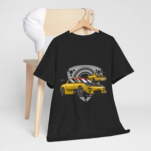 Op de afbeelding: Zwart T-shirt met een gele Mazda RX-7 auto afbeelding. De auto is omringd door een grijs en wit tandwiel en schild ontwerp.