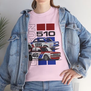 May include: Pink t-shirt featuring a graphic of two vintage Datsun 510 race cars. The cars are white, blue, and red, with the numbers 85 and 46. The word "Datsun" and "510" are printed above the cars in a bold font.