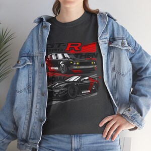 May include: A dark grey t-shirt with a graphic design featuring two black Nissan Skyline GT-R cars. The text "GT-R" is printed in red and white above the cars. The text "THEN NOW" is printed below the cars. The cars are both labeled with "ADVAN" on the side.