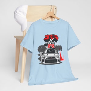 Puede incluir: Camiseta azul claro con un gráfico de Godzilla vs. GT-R. El gráfico presenta un Nissan GT-R blanco con personajes de Godzilla a cada lado.