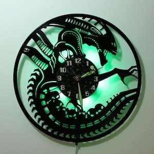 May include: Black vinyl record clock with a green light behind it. The clock face is a black and white design of an alien creature with the numbers 1-12 around the edge.