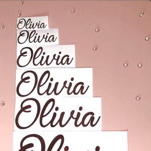 Può includere: Sei adesivi in ​​vinile color oro rosa di diverse dimensioni con il nome "Olivia" scritto in corsivo. Gli adesivi sono su carta bianca e sono disposti in una pila dal più grande al più piccolo.